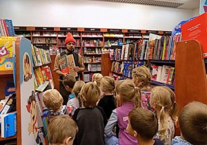 Dzieci stoją między regałami z książkami i patrzą w kierunku mężczyzny w czerwonej czapce krasnala.